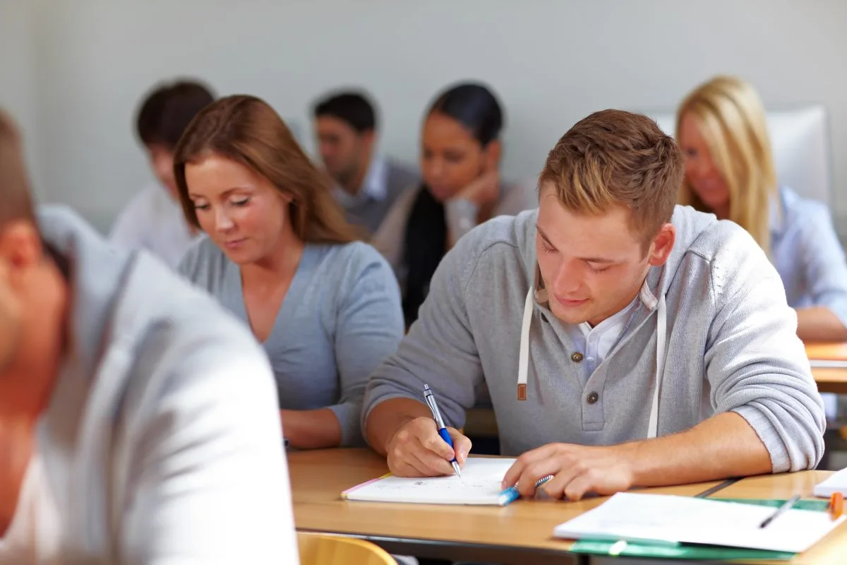 A smiling student in a classroom holding a textbook, with other students in school uniforms sitting in the background